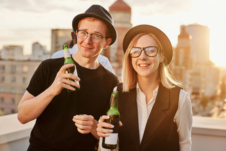 Terrace hangout at sunset. Portrait of young stylish hipster-like Caucasian man and woman standing on rooftop with bottles of beer and smiling at cameraの写真素材