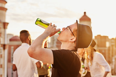 Young ginger man in bowler hat and glasses drinking beer from bottle while hanging out with friends on rooftop terrace on sunny eveningの写真素材