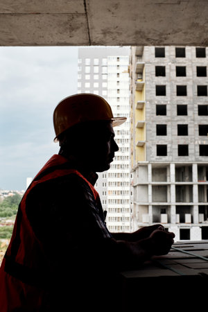 Man wearing construction helmet standing on building site balcony overlooking unfinished high rise building, holding hands together, facing cityscapeの写真素材