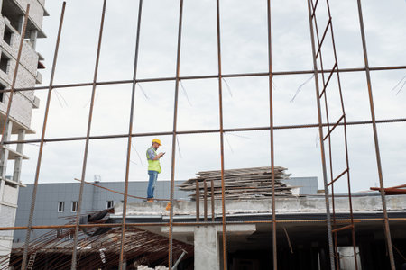 Caucasian middle aged man wearing safety helmet standing on construction site using smartphone, surrounded by steel framework and building materials during workdayの写真素材