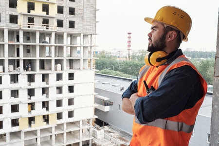 Caucasian man wearing construction helmet and safety vest standing with arms crossed looking at unfinished building, overseeing construction site progressの写真素材