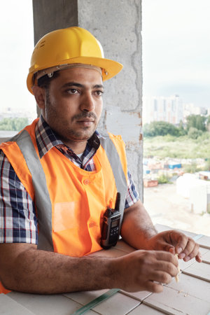 Male builder wearing construction helmet and safety vest standing at building site holding toothpick looking away with serious expressionの写真素材