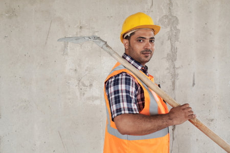 Portrait of young adult man wearing construction helmet and safety vest holding shovel over shoulder...