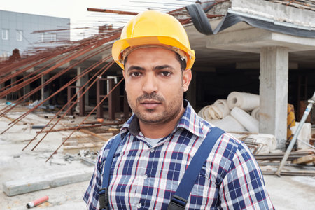 Portrait of young adult Middle Eastern man wearing hard hat standing at construction site looking at camera, facial expression serious, visible building materials and structureの写真素材