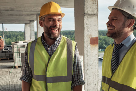 Two male coworkers wearing safety vests and hard hats standing on construction site smiling and...