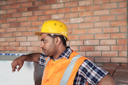 South Asian man wearing safety vest and hard hat standing at construction site leaning on surface looking to side showing serious expressionの写真素材