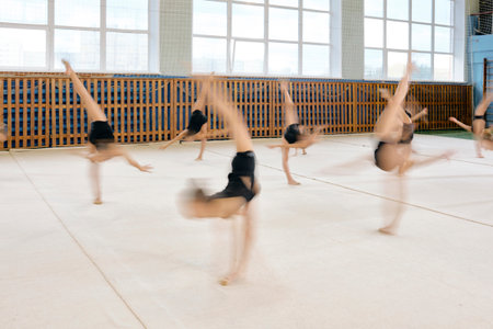 Group of Caucasian teenage girls performing synchronized gymnastics routines on gym floor, bodies in motion with blurred effect, demonstrating athletic skill and coordination indoorsの写真素材