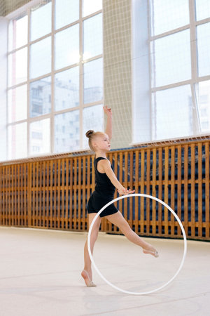 Caucasian girl performing rhythmic gymnastics routine with hoop, standing on one leg and raising arm in sports hall with large windows, demonstrating balance and flexibilityの写真素材