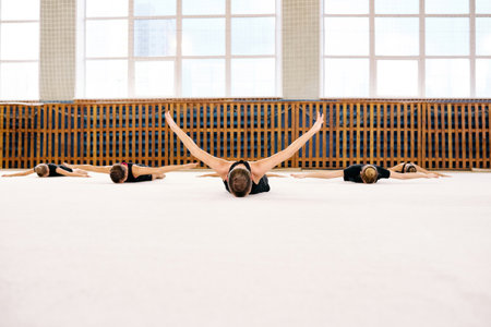 Group of Caucasian teenagers lying on gym floor stretching arms and legs during synchronized gymnastics practice, bodies aligned in formation, large windows in backgroundの写真素材