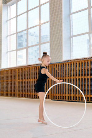 Caucasian girl child practicing rhythmic gymnastics holding hoop with one hand standing on tiptoe in gymnasium, focused expression, hair in bun, large windows in backgroundの写真素材