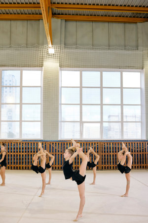 Group of Caucasian girls practicing rhythmic gymnastics in sports hall, each performing standing split pose with one leg raised high, focusing on balance and flexibility trainingの写真素材