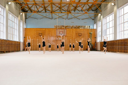 Group of Caucasian girls performing synchronized gymnastics routine in spacious gymnasium, each raising arms above head while standing on mat, wooden sports equipment visible in backgroundの写真素材