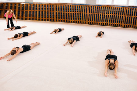 Group of Caucasian girls lying on gym floor stretching arms forward during gymnastics class, female coach supervising from side, all participants wearing athletic outfits, indoor sports facilityの写真素材