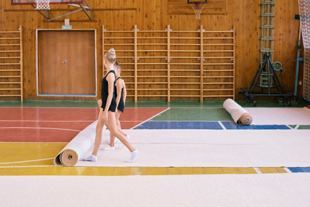 Two girls walking across gymnasium floor wearing black leotards, moving past rolled mats, wooden wall bars and basketball hoops visible in background