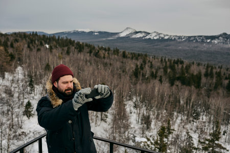 Traveling man in warm outwear and hat using phone and taking photo of landscape from high point in winter timeの写真素材