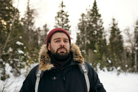 Portrait of adult bearded man in warm jacket and hat standing with backpack in winter woods looking at cameraの写真素材