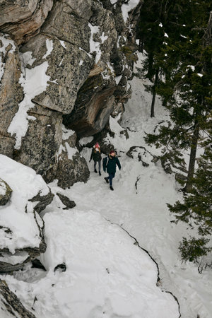From above view of hiking woman and man with backpacks walking alongside cliff in coniferous winter woodsの写真素材