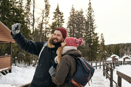 Young happy woman and man with backpack in outwear taking selfie on wooden walkway in snowの写真素材