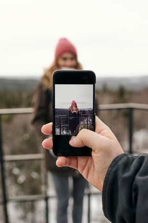 Crop male hand using smartphone and photographing smiling girlfriend on terrace against highlands