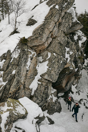 View of big rocky cliff in snowy forest with couple trekking together and walking below
