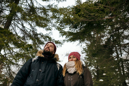 From below of young woman and man in outwear standing in coniferous winter woods looking away dreamilyの写真素材