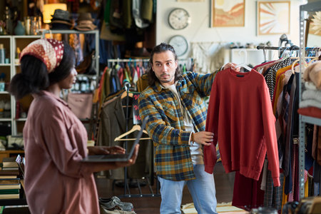 Caucasian young adult man showing sweatshirt to Black young adult woman while shopping in thrift shop, man holding clothing on hanger and woman gesturing with hands, racks of clothes in backgroundの写真素材