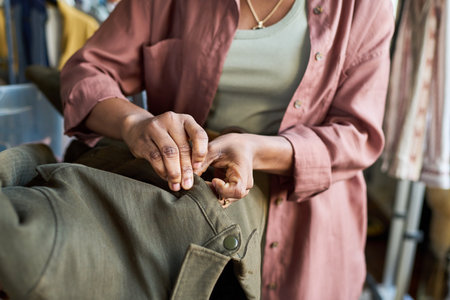 Young adult Black woman adjusting buttons on coat inside thrift shop, hands carefully fastening...