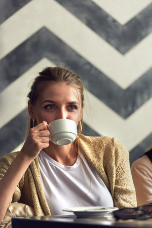 Daydreaming in coffeehouse. Portrait of young attractive blond woman sitting in cafe, drinking coffee and looking away thoughtfullyの写真素材