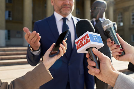 Caucasian middle aged man in suit speaking to group of journalists holding microphones and smartphone outdoors, giving interview or statement in front of government buildingの写真素材