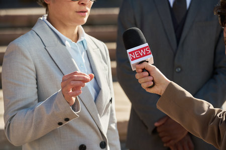 Middle aged Caucasian woman speaking during outdoor interview, gesturing with hand while unidentified journalist holding microphone labeled news, business professional standing nearbyの写真素材