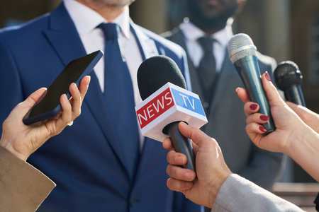 Caucasian middle aged man in suit speaking to group of journalists holding microphones and smartphone during outdoor press conference, hands of reporters visible in foregroundの写真素材
