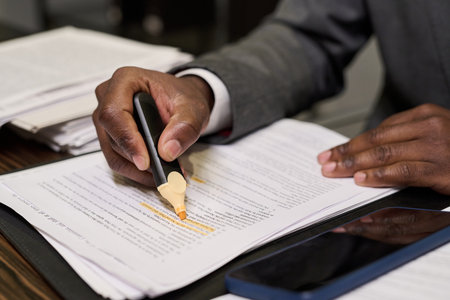Unrecognizable Black man highlighting text on printed documents with highlighter pen, sitting at desk with smartphone nearby, focusing on reviewing paperwork and marking important informationの写真素材
