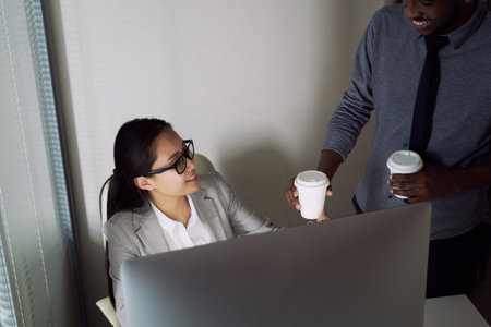 Young African American man bringing coffee to his Asian female colleague sitting at computer in...