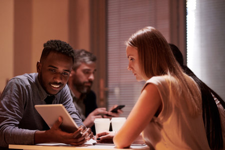 Young African American man showing his Caucasian female colleague something on tablet while sitting in office break room with coffeeの写真素材