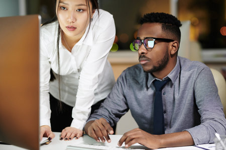 Multiethnic couple of workaholics. Young Asian woman and her male African American colleague looking attentively at computer screen while working late in officeの写真素材