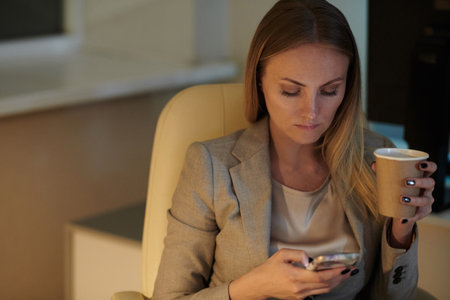 Employee resting in the end of working day. Attractive woman sitting in comfortable office chair, holding cup of coffee and using smartphoneの写真素材