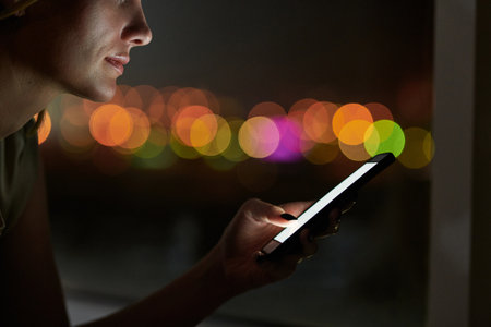 Portrait of businesswoman working till late night. She standing in dark office and texting on smartphone on background of blurred lightsの写真素材