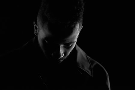 Black and white portrait of African American guy in leather jacket standing in the dark alone and looking down sadlyの写真素材