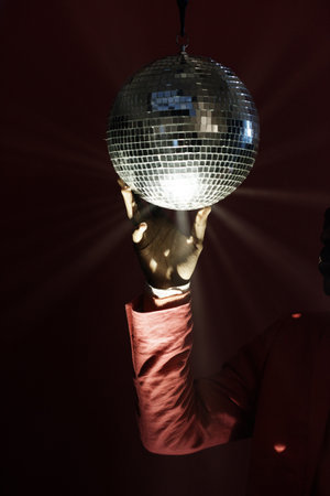 Symbol of 80s party. Unrecognizable man in red suit jacket rotating shiny disco ball in retro nightclub in darknessの写真素材