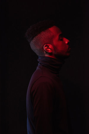 Profile portrait of young handsome African American man with stylish haircut standing in the dark with his eyes closedの写真素材