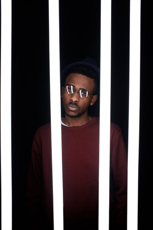 Conceptual portrait of young fashionable Black man in glasses standing behind luminescent lamps in the darkの写真素材