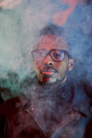 Portrait of stylish African American guy in leather jacket and glasses standing in cloud of smoke and looking at cameraの写真素材