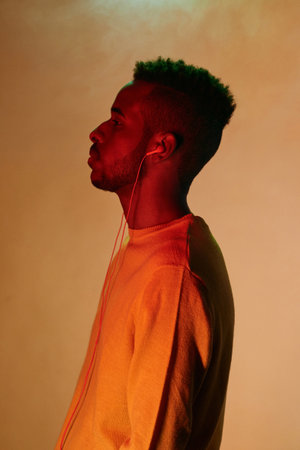 Profile portrait of young stylish Black guy with earphones standing and looking into the distance while listening to musicの写真素材