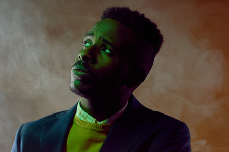 Portrait of young fashionable African American guy standing in the dark surrounded by smoke and...