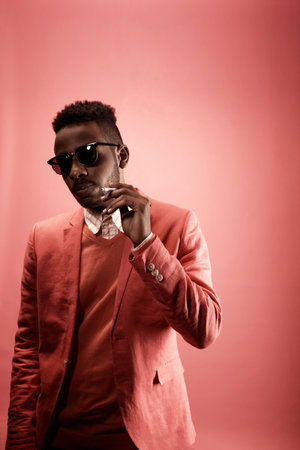 Portrait of young stylish Black man in sunglasses looking at camera while standing against pink...