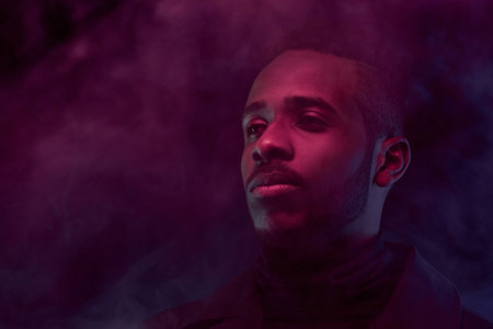 Portrait of young attractive Black man standing in purple smoke cloud in the dark and looking into...