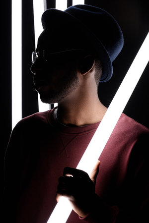 Waist-up portrait of stylish Black man in hat and glasses standing in the dark with fluorescent lamp...