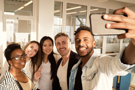 Multiethnic group of young colleagues smiling happily at smartphone camera while taking selfie in modern officeの写真素材