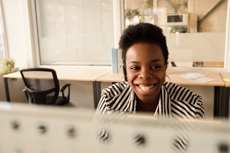 Portrait of stylish black woman looking away and smiling cheerfully while sitting at table in modern office with partition wallsの写真素材