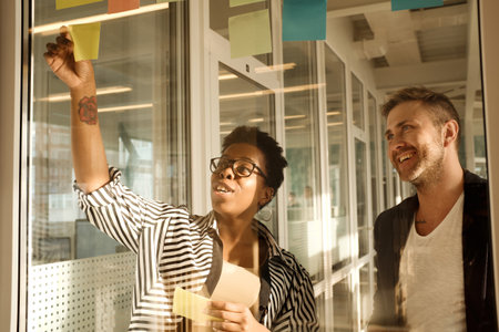 Stylish black woman and her caucasian male coworker looking at sticky notes on glass wall in office...